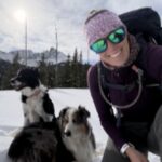 female wearing sunglasses and a knit hat standing in the snow with 2 dogs